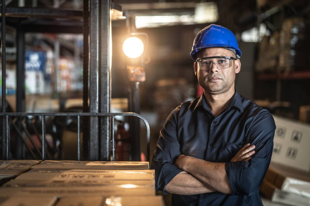 Male worker in warehouse standing front on with arms crossed wearing a protective helmet and glasses