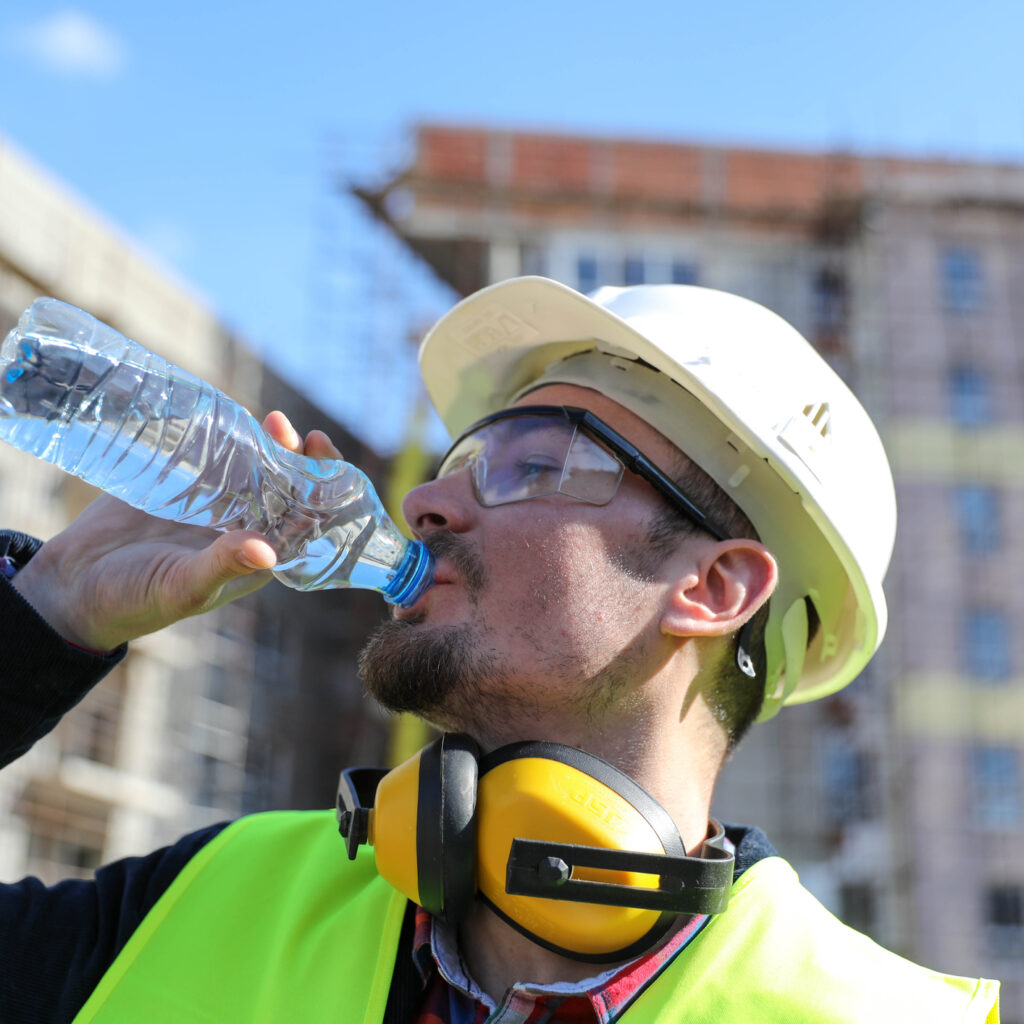 Male tradesman working outdoors in high-vis vest and protective gear drinking bottle of water to protect himself from heat injury