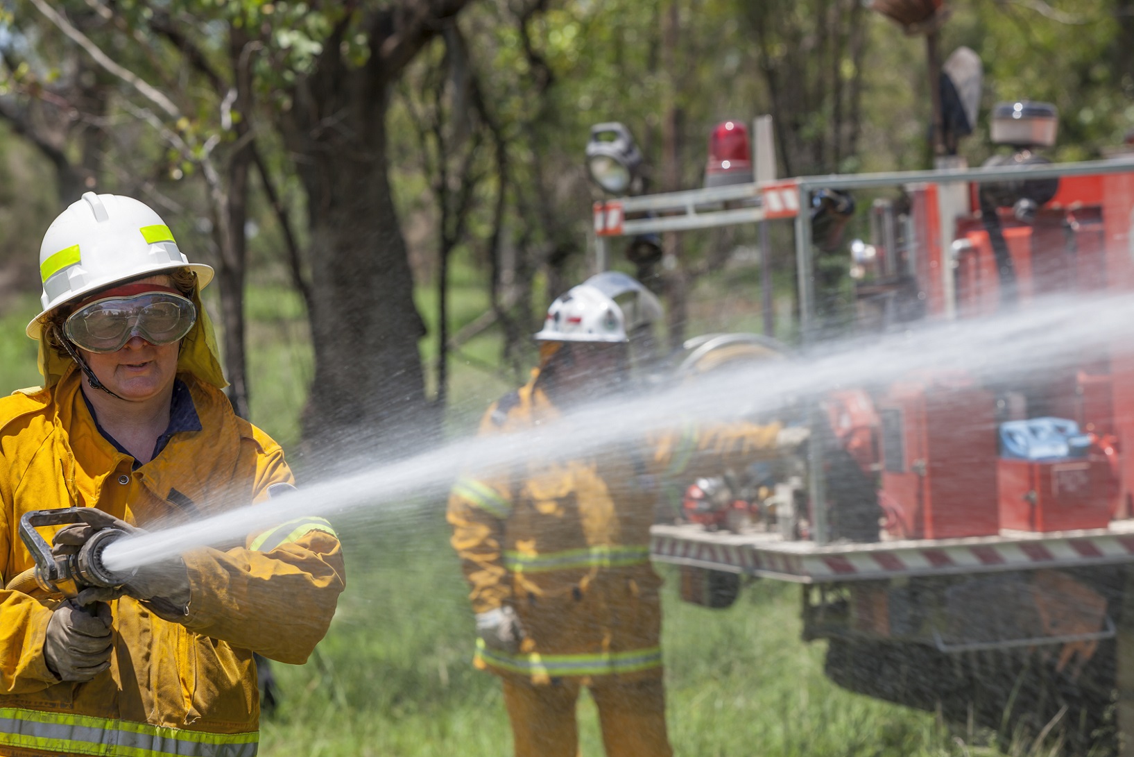 A female rural fire fighter with hose and fire truck. She is dressed in protective clothing.